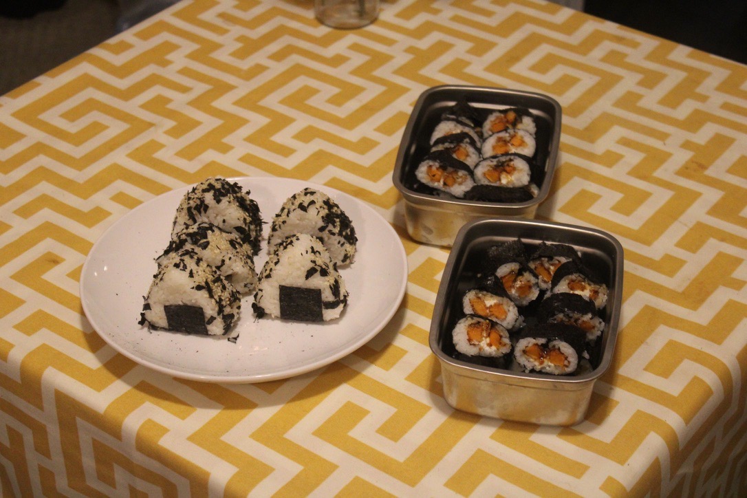 a plate with five onigiri, as well as two metal containers each with 16 sweet potato sushi rolls 
